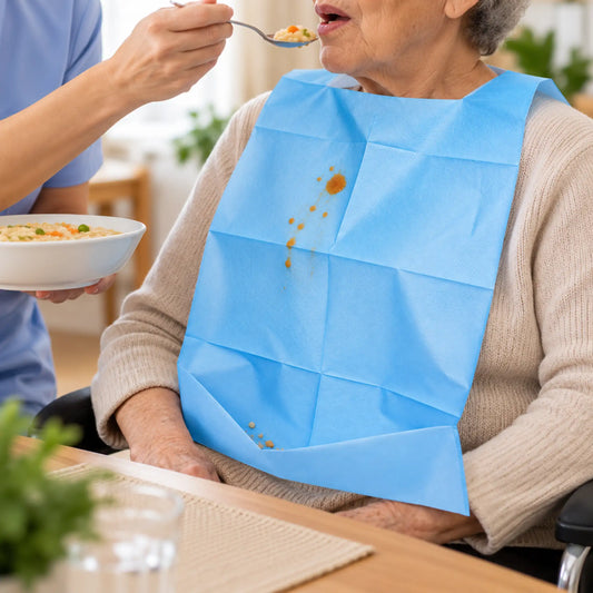 Persona mayor en silla de ruedas utilizando un babero de papel con bolsillo mientras recibe asistencia durante la comida.