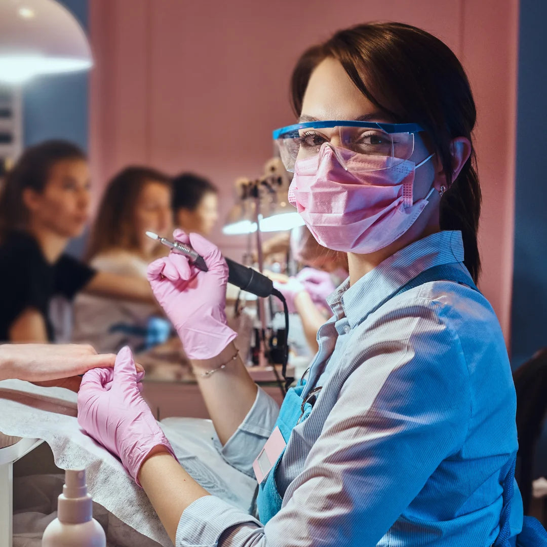 Manicurista con mascarilla quirúrgica rosa Tipo IIR y guantes rosas durante un servicio profesional de manicura